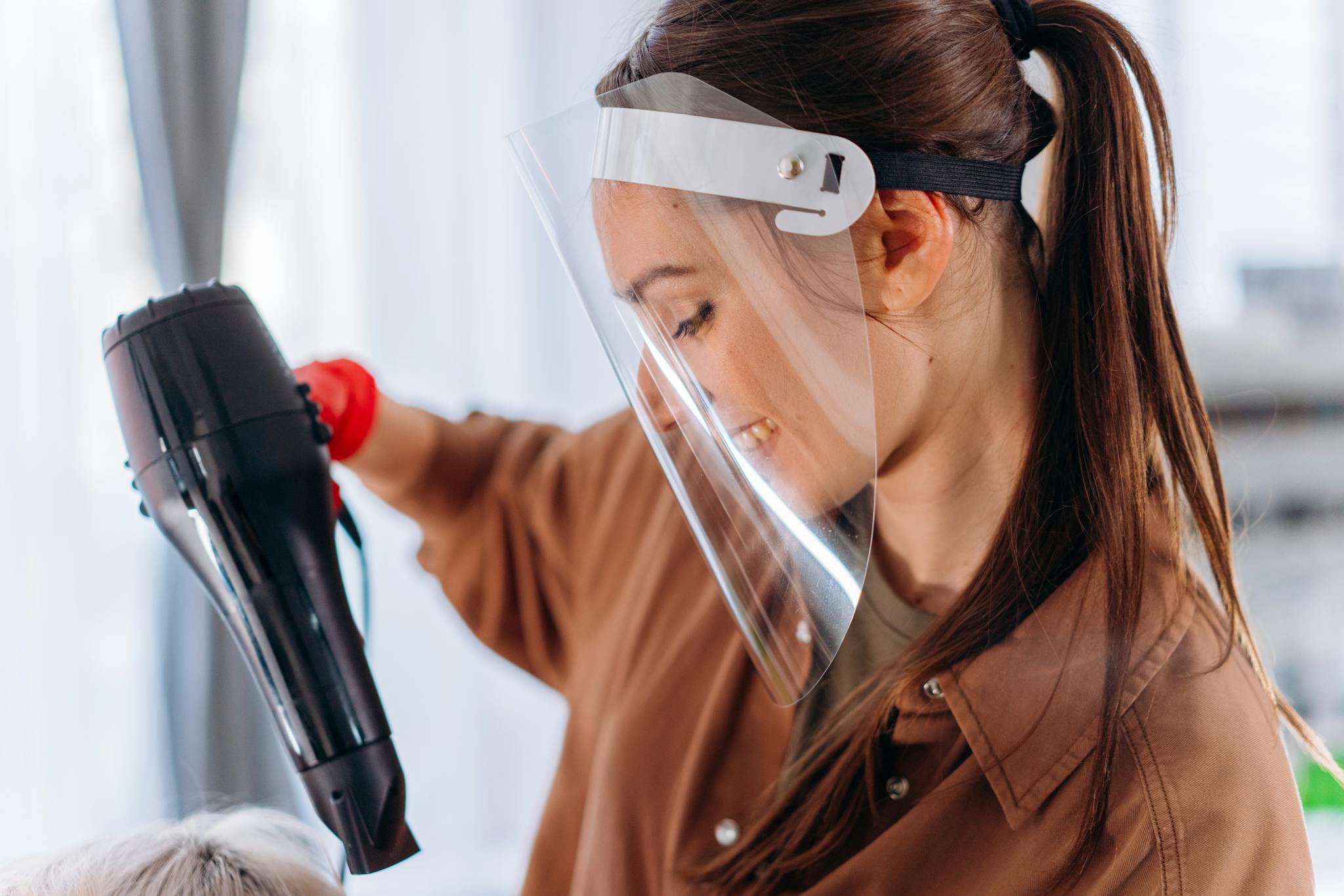 Hairdresser wearing a face shield, using a hair dryer to style hair. Indoor salon setting with a safety focus.
